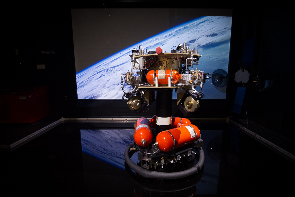 The ASTROS platform with orange propellant tanks on its air-bearing pedestal, Earth projected behind