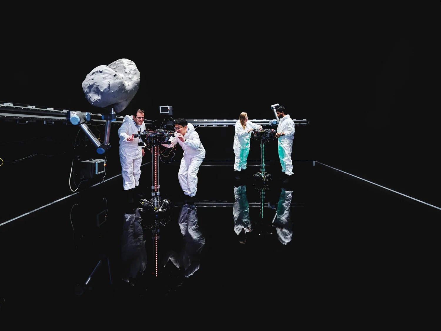 Four researchers in white suits tend two pedestal robots on a glossy black floor, an asteroid model on a rail behind