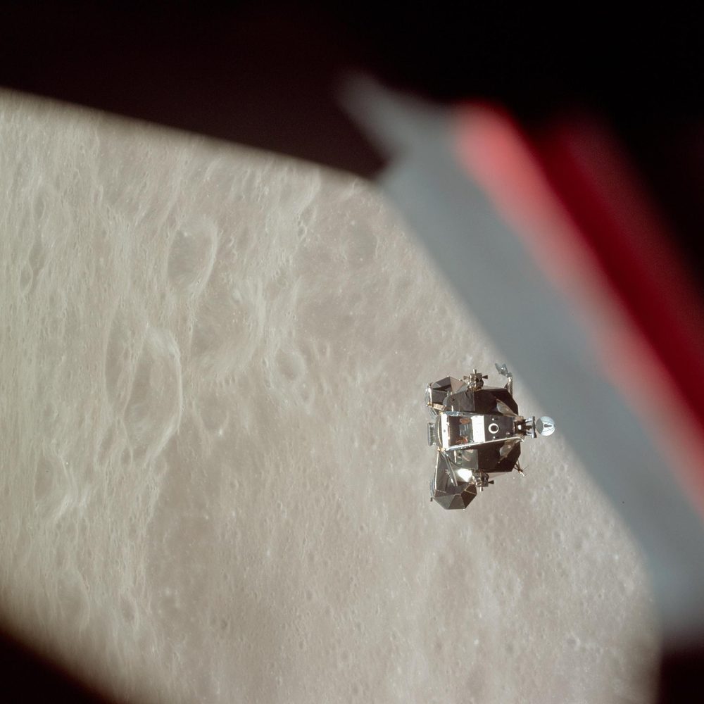 Small bright vehicle against the grey cratered Moon, seen past a window frame edge