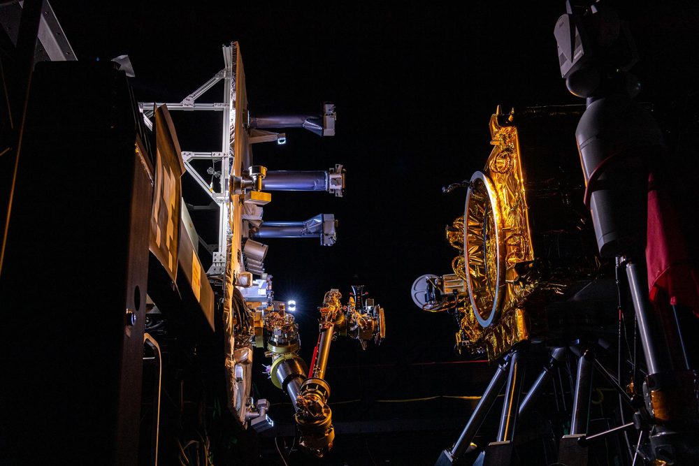 A robotic arm reaches toward a gold-foil satellite mock-up with a dish, in near darkness