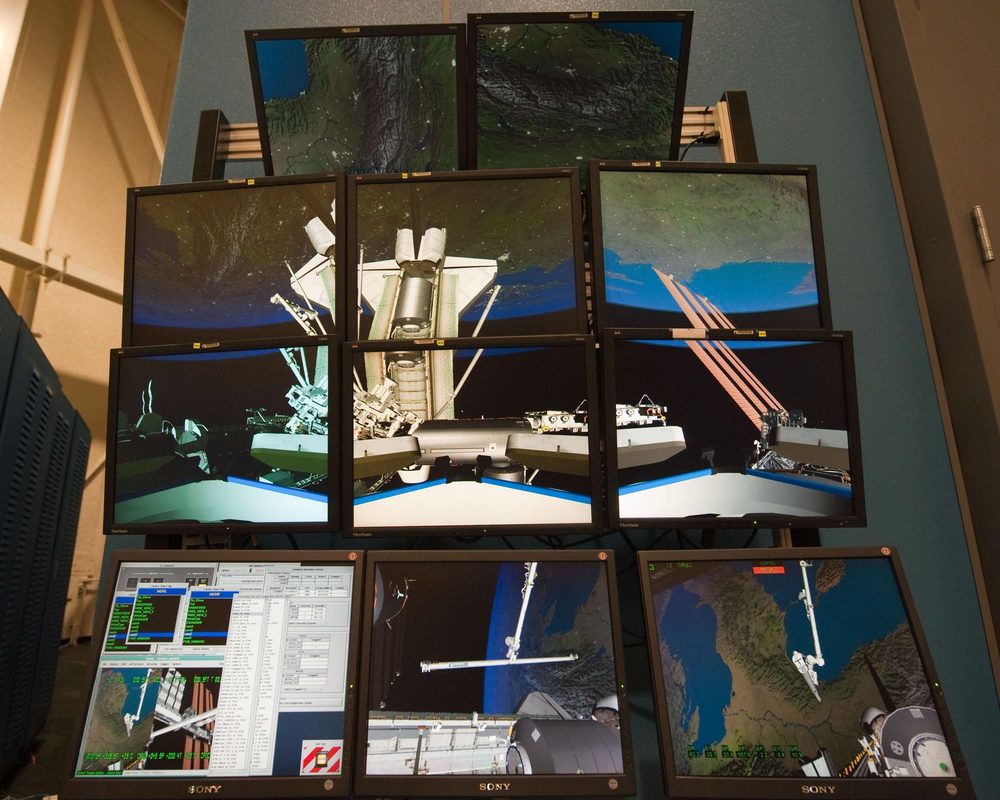 Bank of stacked flat monitors showing rendered station structure against Earth, one panel of readouts