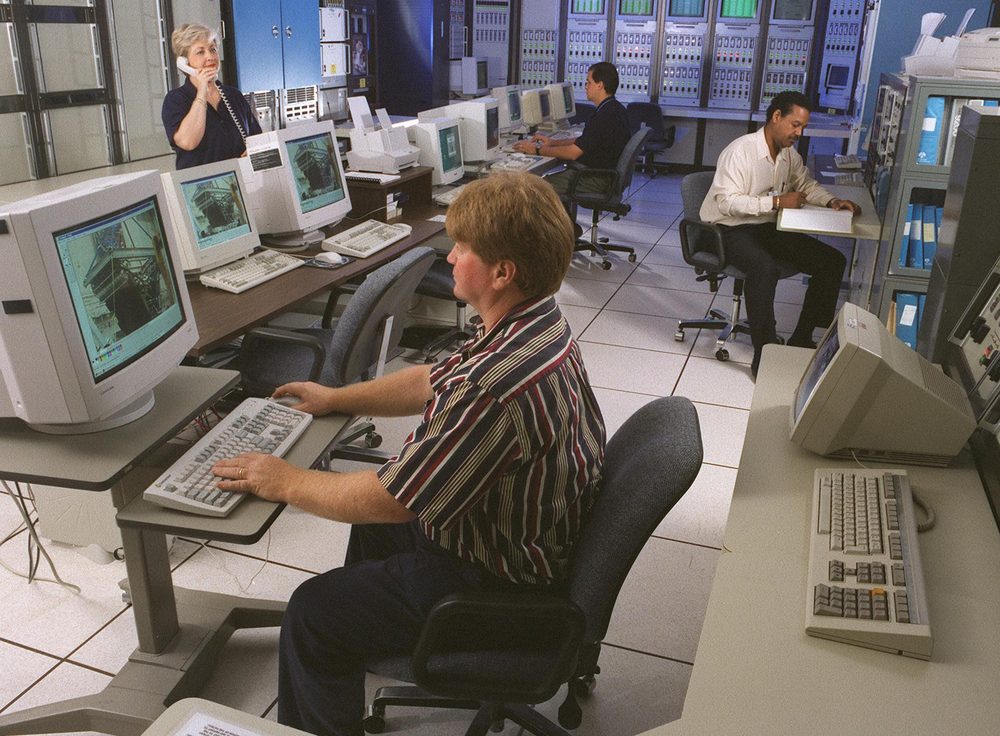 Beige CRT workstations on a raised floor, staff at keyboards, instrument racks along the wall