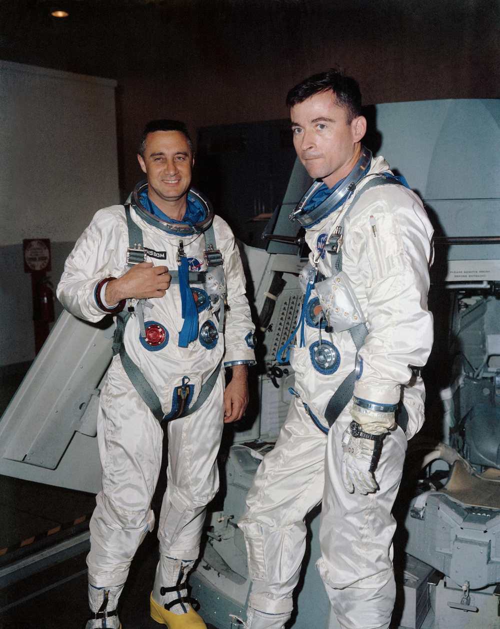 Two men in white pressure suits stand helmetless beside a spacecraft trainer indoors