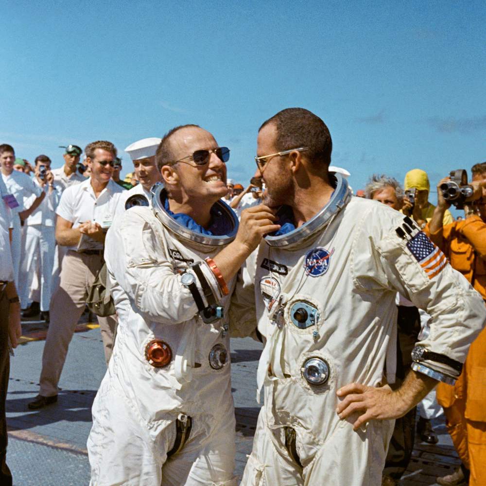 Two crewmen in white suits and sunglasses grin on deck amid photographers and sailors