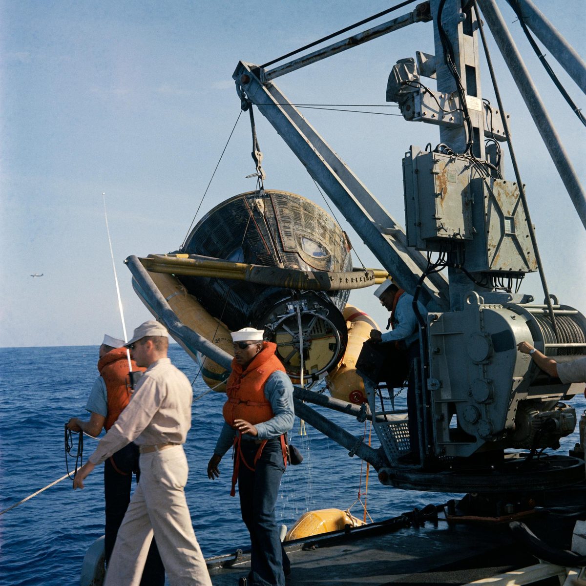 Sailors in life jackets steady a scorched capsule swinging from a shipboard crane