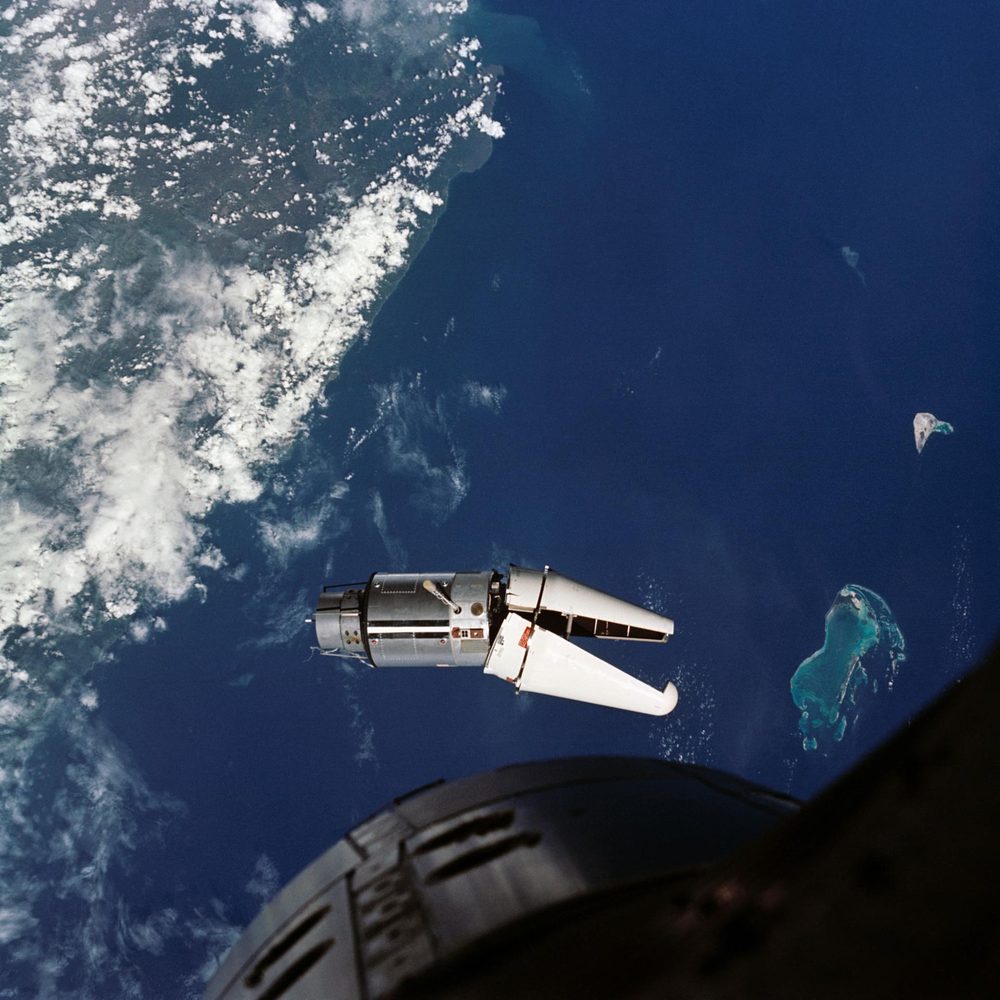 A small silver vehicle above deep blue ocean and a pale reef, spacecraft nose below