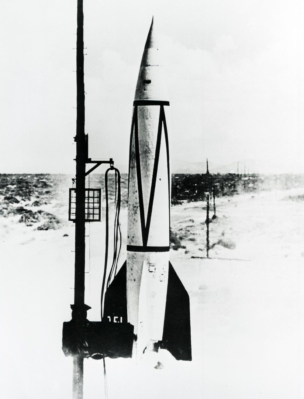 Finned rocket rising past a tall lattice mast over flat scrubby desert, monochrome