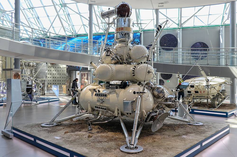 Tall four-legged lander crowded with spherical tanks, on a regolith plinth under a glass roof