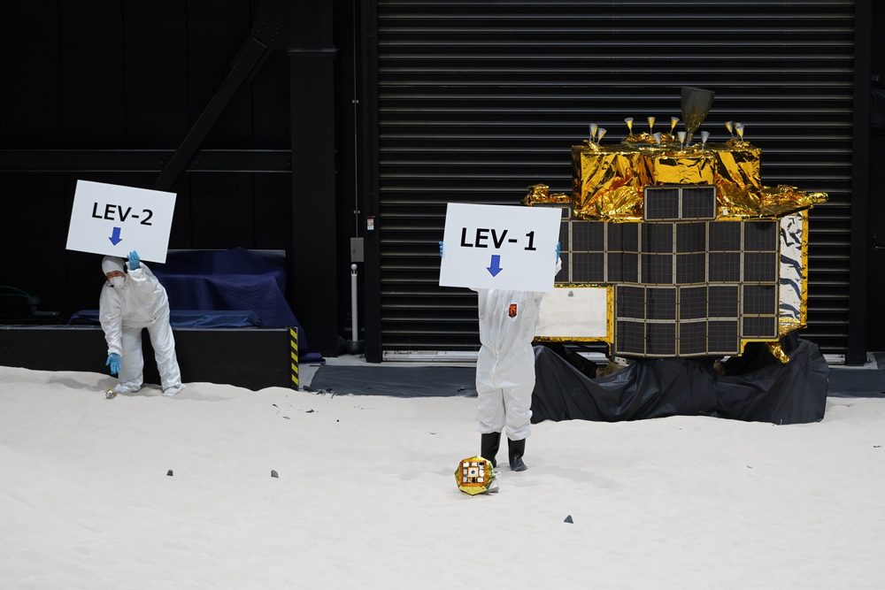 People in white coveralls hold LEV-1 and LEV-2 signs beside a gold-foiled lander