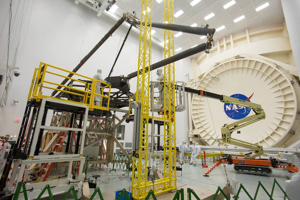 Technicians in cleanroom suits on yellow lifts around black booms, huge chamber door behind