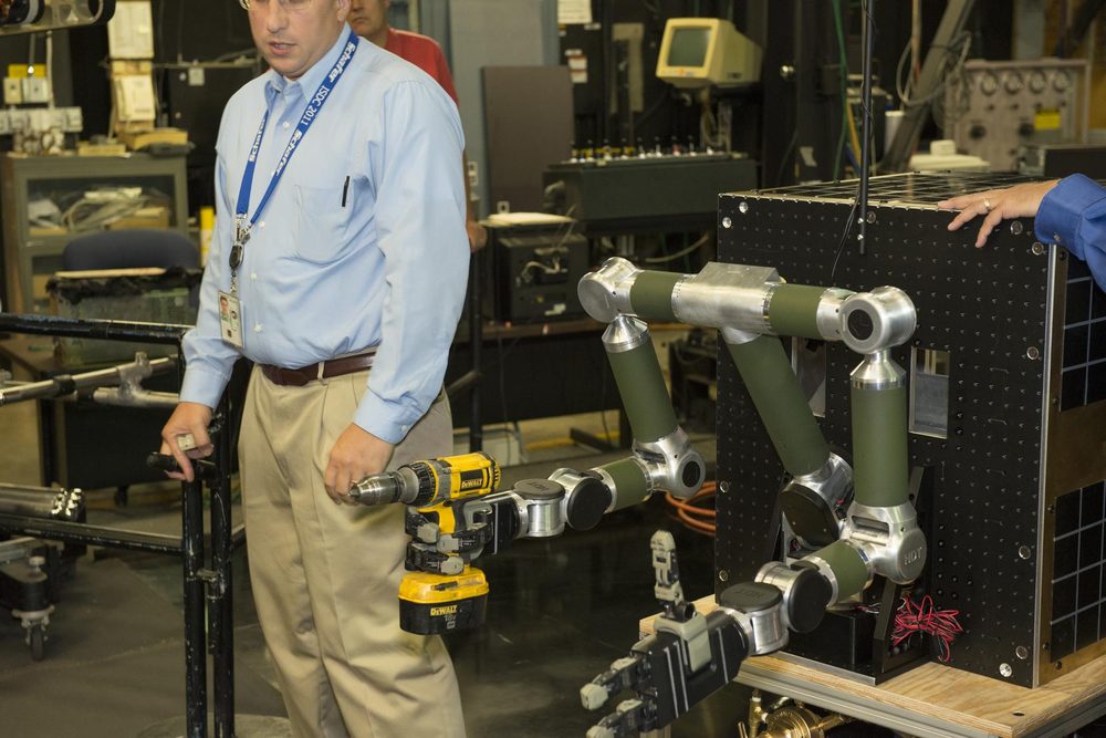 An engineer watches a jointed arm holding a cordless drill against a black panel