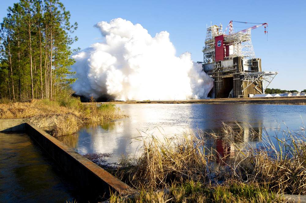 Tall test stand venting a huge white cloud sideways above a still pond and pines